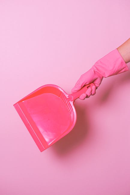 A person wearing a pink rubber glove holding a bright pink dustpan against a solid pink background. The dustpan is made of plastic with a textured handle and a wide, flat lip for collecting debris. The scene emphasizes cleaning tools used for surface cleaning and debris collection, relevant to domestic or commercial cleaning practices. This image is associated with Carpet Cleaning Bromley’s emphasis on hygiene, surface maintenance, and deep cleaning services, as indicated on the webpage about avoiding hidden charges in Bromley cleaning quotes at https://carpetcleaningbromley.net.