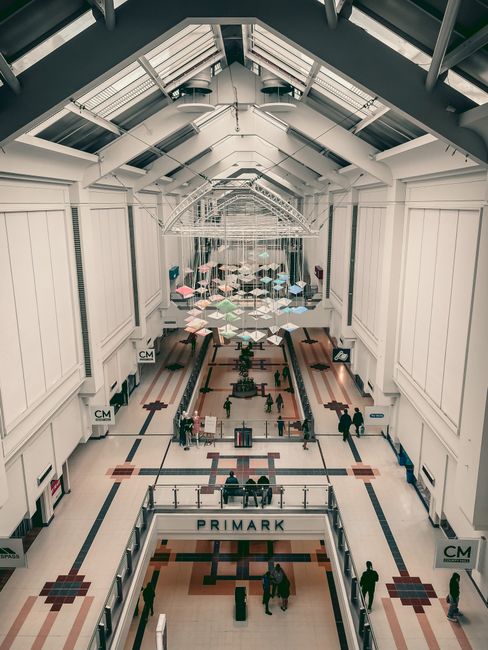 A spacious retail shopping area inside The Glades shops in Bromley, featuring a high vaulted ceiling with large skylights allowing natural light to illuminate the white-paneled walls and tiled flooring. The floor showcases a geometric pattern in beige, black, and red tiles, with a clear, polished surface indicating recent deep cleaning and thorough surface sanitisation. Several shoppers are walking through the corridor, some engaged with digital kiosks or browsing stores. The scene includes modern pendant lighting fixtures hanging from the ceiling, and umbrellas arranged decoratively towards the back of the space. Carpet Cleaning Bromley offers professional cleaning services for commercial interiors like this, ensuring hygiene, surface cleaning, and maintenance of high-traffic shopping environments.