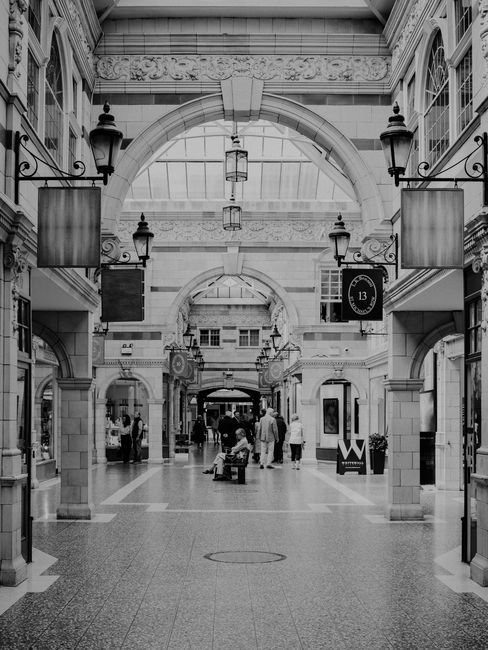 The image depicts an indoor shopping arcade with high vaulted ceilings and ornate architectural details. Large arched openings frame the walkway, which is lined with display windows and storefronts. The polished tiled floor appears clean and well-maintained, reflecting the ambient lighting from decorative lantern-style light fixtures hanging from the ceiling. Several shoppers are walking through the space, some carrying bags, while a person is sitting on a bench in the center. The overall appearance suggests a tidy and hygienic environment, maintained through professional cleaning services. Carpet Cleaning Bromley specializes in surface cleaning and deep cleaning, ensuring commercial spaces like The Glades Shops remain presentable and hygienic for visitors. The scene showcases a bright, open area with a sophisticated aesthetic, emphasizing the importance of regular sanitization and maintenance in such high-traffic environments.