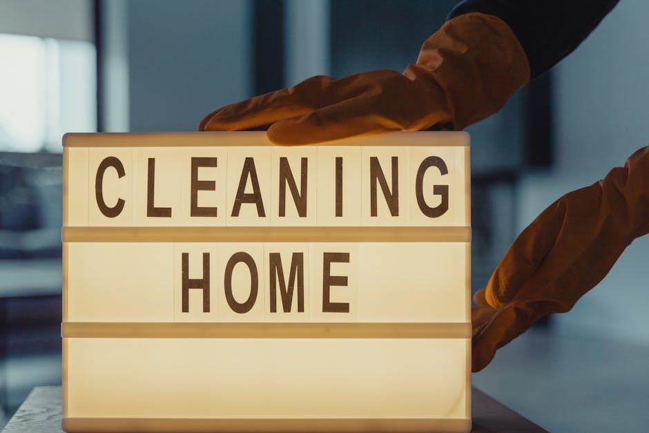 A person wearing brown cleaning gloves is adjusting a backlit sign displaying the words 'CLEANING HOME' in capital letters. The sign is placed on a flat surface in a well-lit room, with a blurred background showing modern interior elements such as windows and furnishings. The surfaces around the sign are clean, suggesting a professional approach to residential or commercial surface cleaning and deep cleaning, as promoted by Carpet Cleaning Bromley in their end of tenancy cleaning success story in Bromley North.