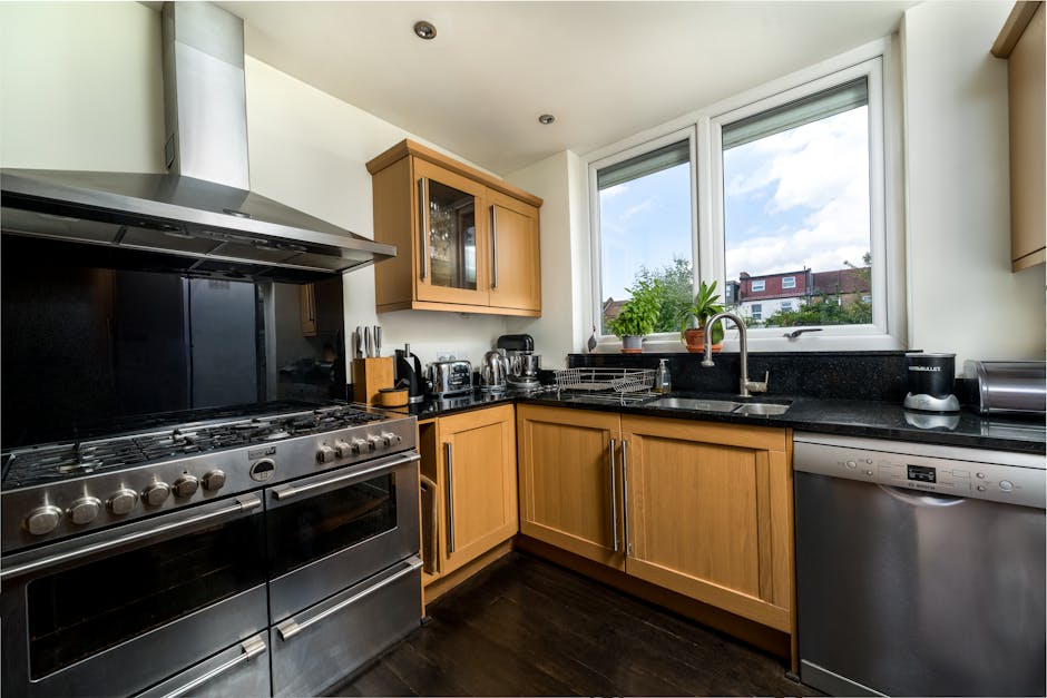 A tidy and well-maintained kitchen featuring light wooden cabinets with stainless steel handles, a black granite countertop, and modern appliances including a stainless steel oven and a dishwasher. The black tiled backsplash contrasts with the white walls, and a large window above the sink allows natural light to brighten the space, revealing a view of residential houses and a blue sky with scattered clouds. On the countertop, there are small kitchen appliances such as a toaster and a kettle, with a potted plant near the window adding a touch of greenery. The surfaces appear spotless and clean, indicative of professional deep cleaning and surface sanitisation services by Carpet Cleaning Bromley, with the room reflecting a high standard of domestic cleaning aimed at maintaining hygiene and freshness.