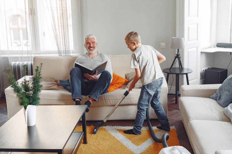 A young boy with blond hair, wearing a beige t-shirt and blue jeans, using a vacuum cleaner to clean a beige sofa in a well-lit living room. An older man with gray hair and a beard, dressed in a light gray shirt and dark jeans, sits on the sofa holding an open book and smiling. The room features a white wall with large windows allowing natural light, a small black side table with a white lamp, a white armchair, a round black table with a potted plant, a colorful orange and white patterned rug, and wooden flooring, all appearing clean and tidy. The scene depicts domestic cleaning and surface maintenance for a hygienic living environment, with Carpet Cleaning Bromley specializing in deep cleaning and sanitisation services.