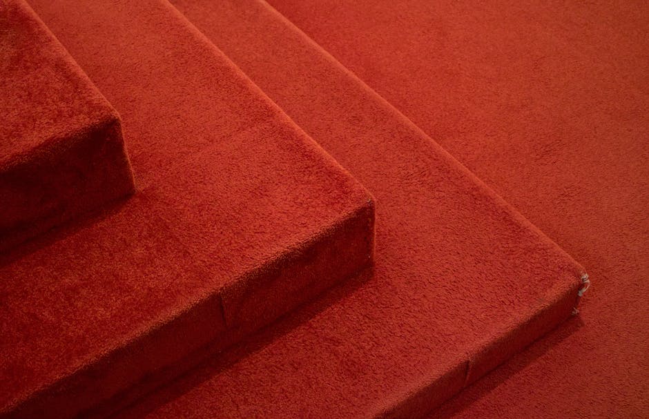 A person wearing a beige shirt is rolling up a light brown, textured carpet from a wooden floor, preparing for removal or deep cleaning. The carpet has a woven pattern, and the surface of the floor un